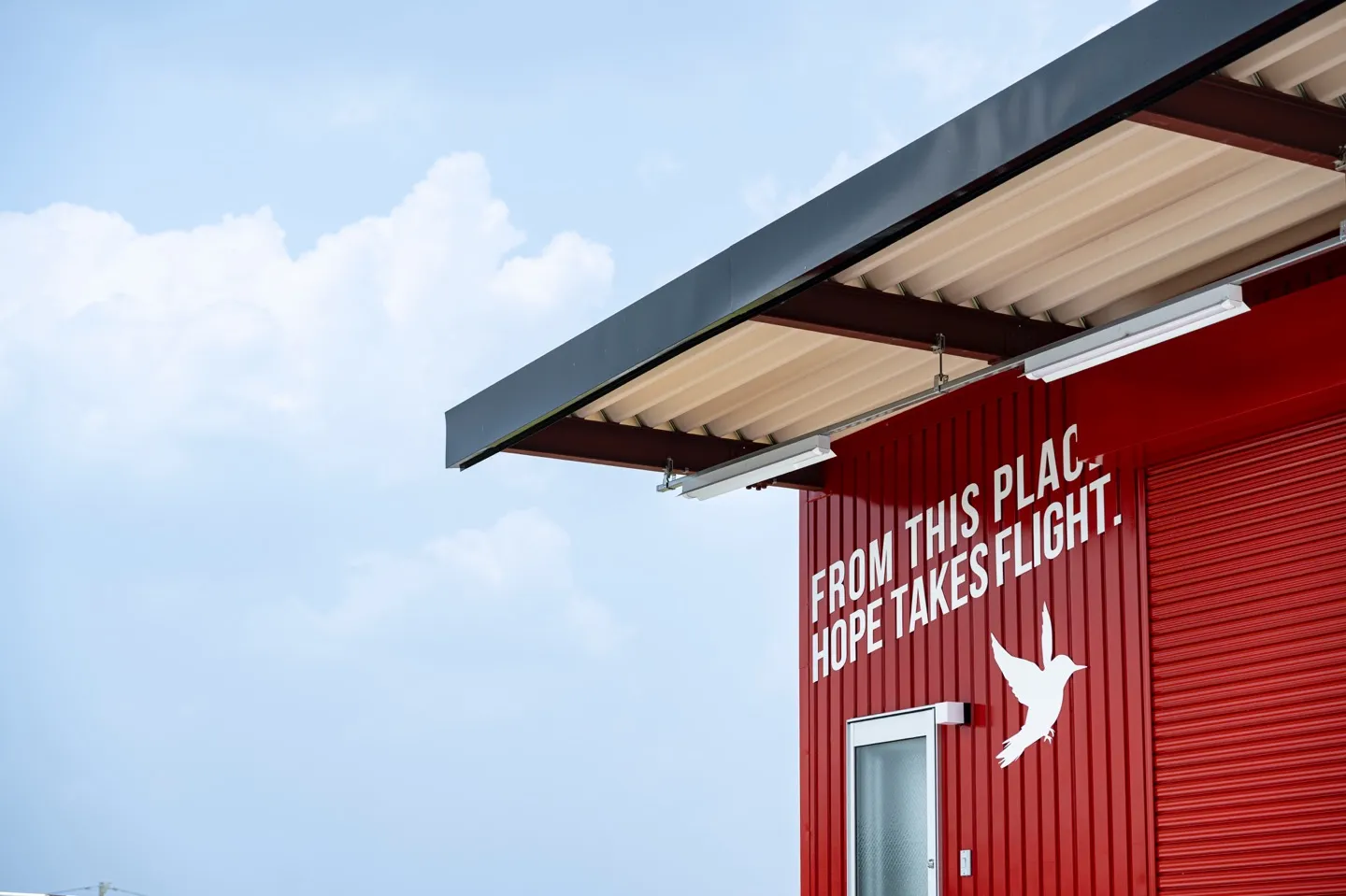 黒瀬ペットフード新社屋の入口側。扉の上に白で「FROM THIS PLACE, HOPE TAKES FLIGHT.」の文字と飛ぶ鳥のシルエット、庇と青空