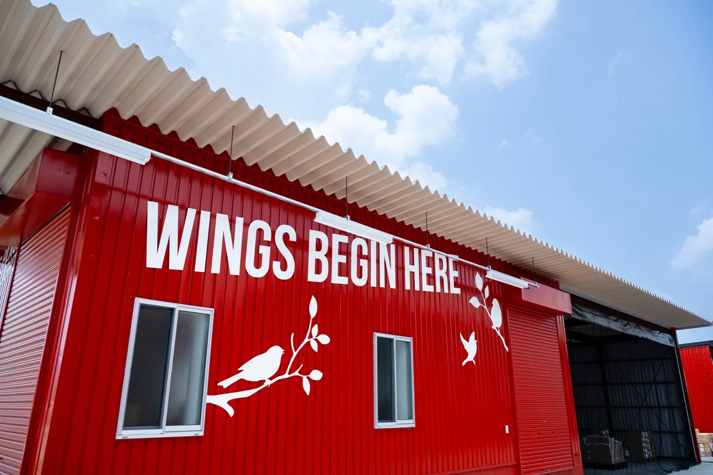 黒瀬ペットフード新社屋の赤い壁面に白で描かれた「WINGS BEGIN HERE」の文字と鳥のシルエット。見上げるアングルで青空が広がる